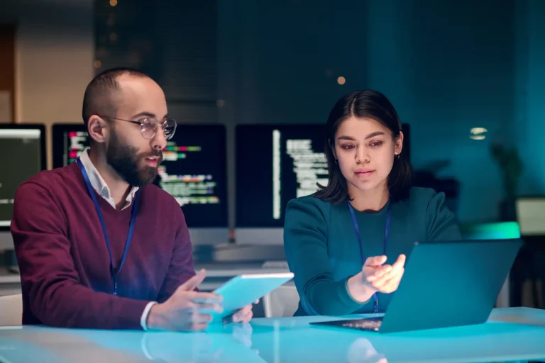 Two colleagues reviewing plans on a laptop and tablet in a shared workspace