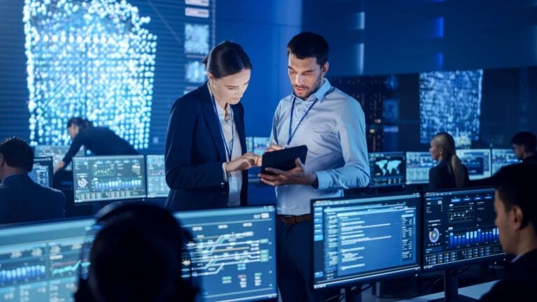 Two colleagues reviewing a tablet in a security operations centre while analysts monitor dashboards