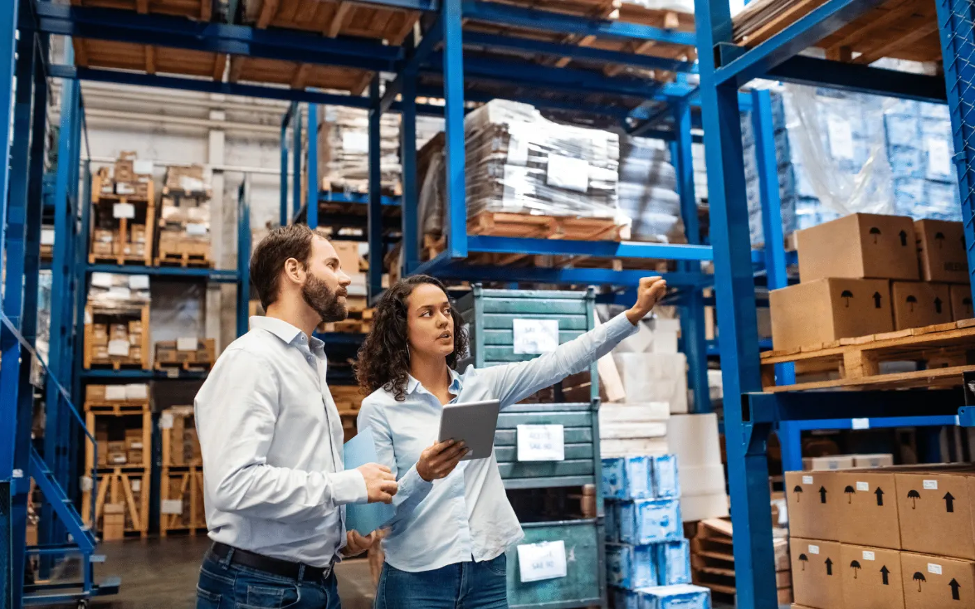 Two staff checking boxed equipment on warehouse racking, one holding a tablet